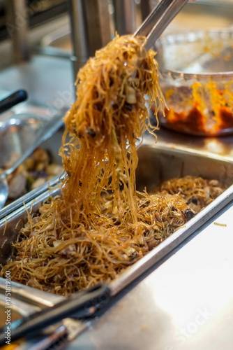 A close-up of a hand using tongs to serve fried vermicelli noodles from a stainless buffet tray in an Indonesian food stall. Authentic Asian street food scene with warm lighting and real-life