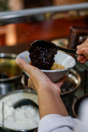 Hand serving traditional Indonesian black glutinous rice porridge, known as bubur ketan hitam, into a bowl with coconut milk. Authentic Asian dessert scene showing local food culture