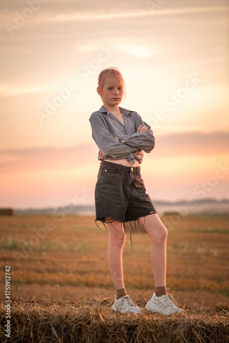 Young woman in casual attire stands in a field at sunset, surrounded by hay bales, showcasing a serene atmosphere and natural beauty with vibrant colors