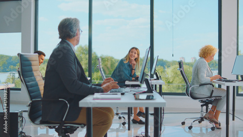 Diverse business professionals work at individual computer workstations in a bright modern office with large windows overlooking lush greenery, fostering collaboration and productivity