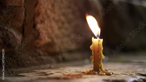 A single, lit yellow candle with dripping wax stands on a textured surface in a dimly lit, rustic setting, casting a warm glow.