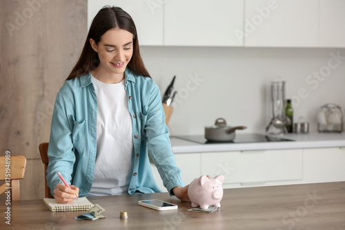 Young woman with piggy bank writing on notebook in kitchen
