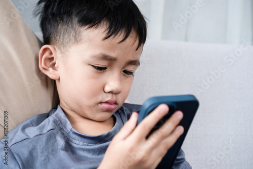 Photos little Asian boy sitting on the sofa and staring at a mobile phone screen, showing signs of smartphone addiction and eye problem
