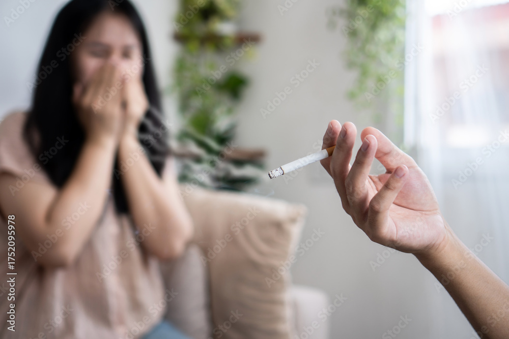 Obraz premium An Asian husband hand smokes next to his wife, who covers her nose to avoid the smell of cigarette smoke, demonstrating the harmful effects of secondhand smoke and bad habits at home.
