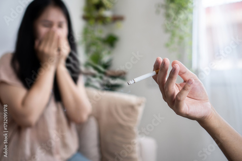 An Asian husband hand smokes next to his wife, who covers her nose to avoid the smell of cigarette smoke, demonstrating the harmful effects of secondhand smoke and bad habits at home.