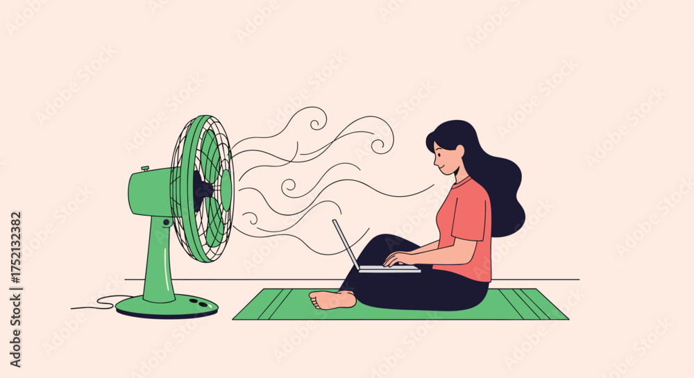 Fototapeta premium A woman sits on a mat working on her laptop while a fan blows air on her to cool her down.