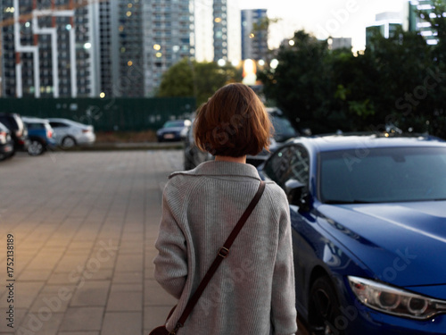 A person stands with a shoulder bag in a paved parking area, facing away from the camera. Urban skyline and parked cars compose a calm, modern city scene.