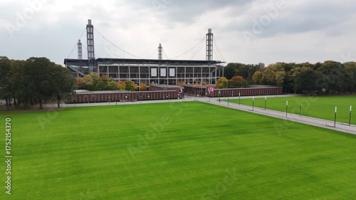 Cologne, Germany - 12 October, 2025: Wide lawn in front of a modern football stadium in Muengersdorf under cloudy sky. Long facade with brick pavilions and trees. Urban park sports venue. No people.