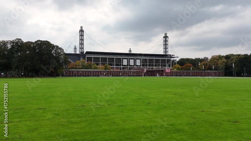 Cologne, Germany - 12 October, 2025: Wide lawn in front of a modern football stadium in Muengersdorf under cloudy sky. Long facade with brick pavilions and trees. Urban park sports venue. No people.