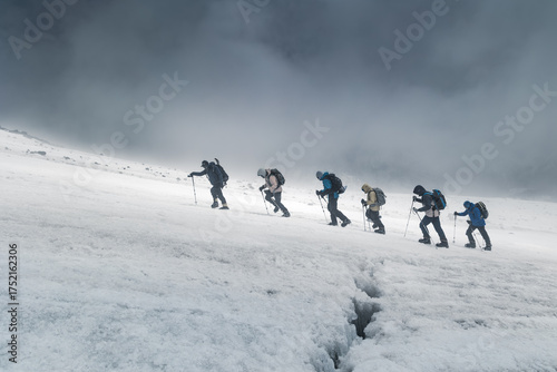 Group of climbers follow instructor walking up cracked icy mountain slope on cloudy day, Elbrus