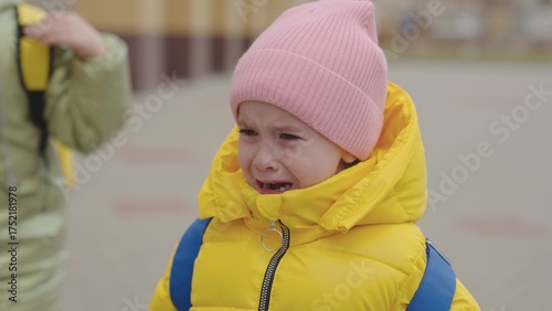 a little child with a backpack is crying in his mother's arms, tears are rolling down kid's face, little girl is upset, daughter is complaining to her mother, schoolgirl is at school with her parent.