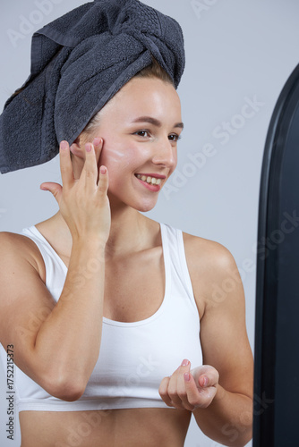 A young beautiful girl in white lingerie with a towel on her head stands in front of the mirror and does cosmetic procedures.