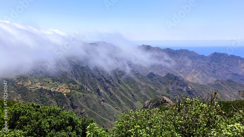 Mountain landscape of Anaga Rural Park, Tenerife. Rolling clouds descending over green valleys and terraced fields with the Atlantic Ocean in the distance under bright midday light.