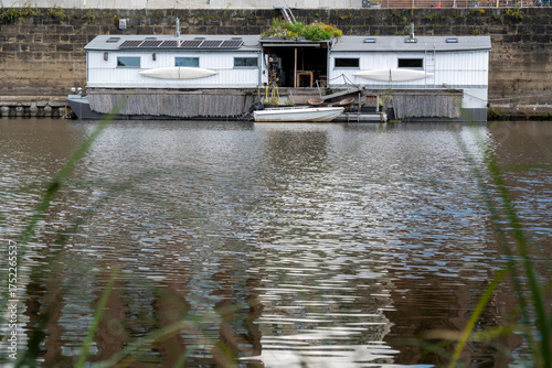 Modernes, weißes Hausboot mit Solaranlage, kleiner Terrasse und Beiboot auf ruhigem Wasser, umgeben von städtischer Uferbefestigung. Wohnen auf dem Wasser.