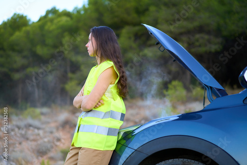 Girl in reflective vest stands by her broken car on a quiet road surrounded by trees during daytime