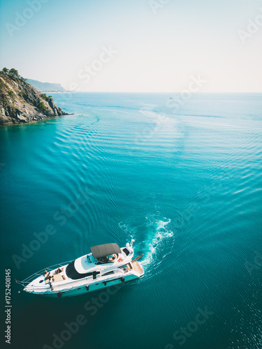 Drone view of a white luxury yacht cruising in turquoise sea near rocky coast. Sunny weather, clear water, and vacation vibes.