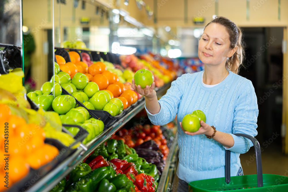 Obraz premium Delighted middle-aged woman customer buying fresh apples at the counter in big greengrocery
