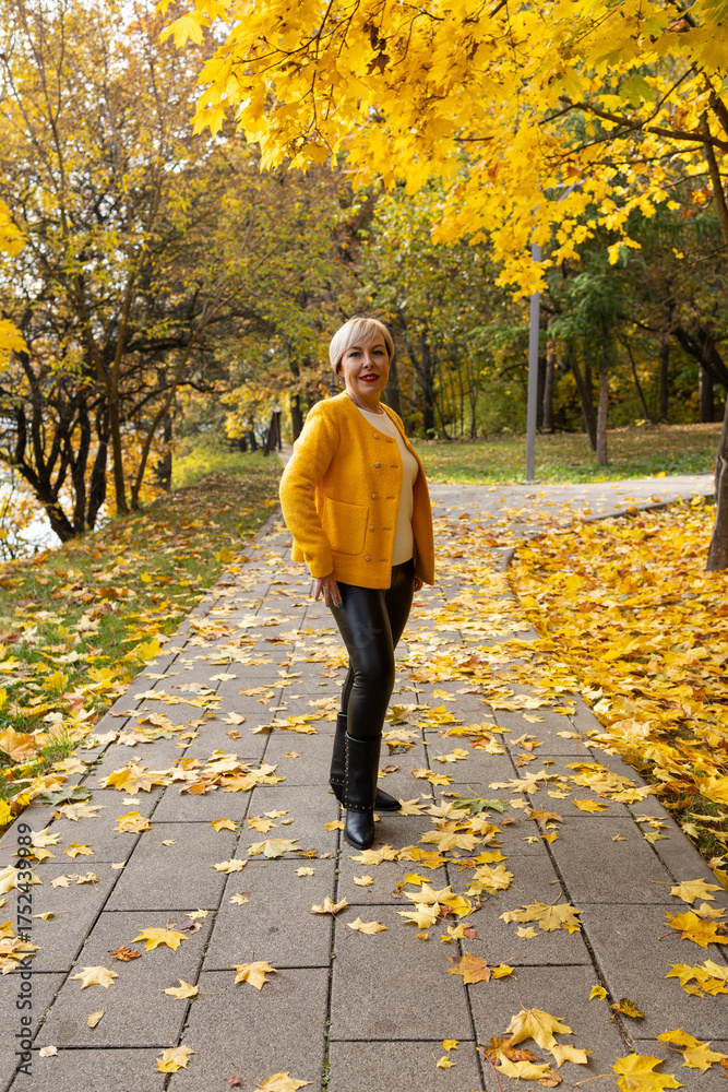 Fototapeta premium woman with short blonde hair stands in a park surrounded by autumn leaves. She wears a bright yellow jacket and black pants.