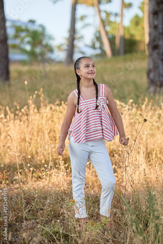 Young girl enjoying a sunny day in a field surrounded by trees and tall grass while wearing a striped top and white pants