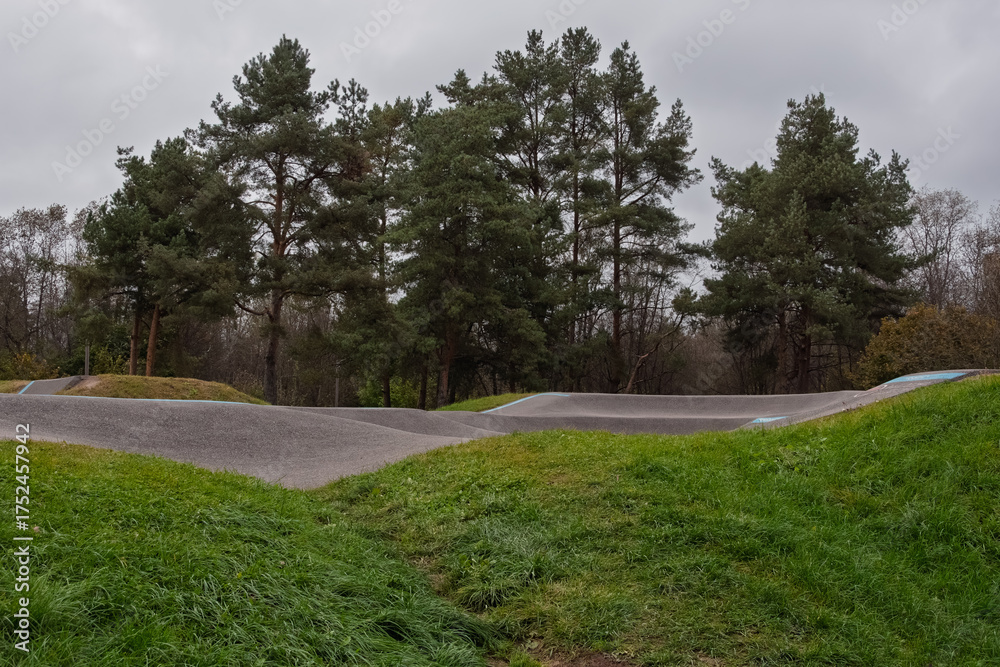 Fototapeta premium A modern asphalt pump track (or skate park) with flowing asphalt waves, set against a backdrop of dark pine trees under a gray, overcast sky. Narva, Estonia.