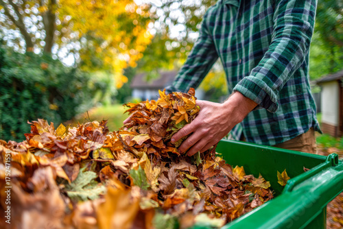 Person gathering and disposing colorful dry autumn leaves into a green garden waste bin on a sunny fall day with blurred natural background