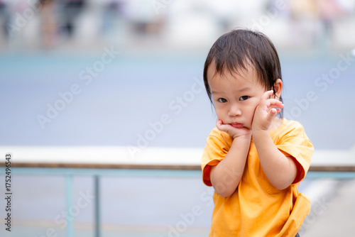 A young Asian boy in a yellow shirt stands outdoors with a blurred background, holding his cheeks with a sad or pensive expression. The photo focuses on his emotional state. Copy space.