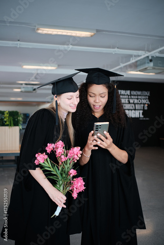 Two diverse female graduate students, teenage friends watching celebration photos on mobile phone together, degree achievement milestones, happiness in academic college, and cheerful congratulations.
