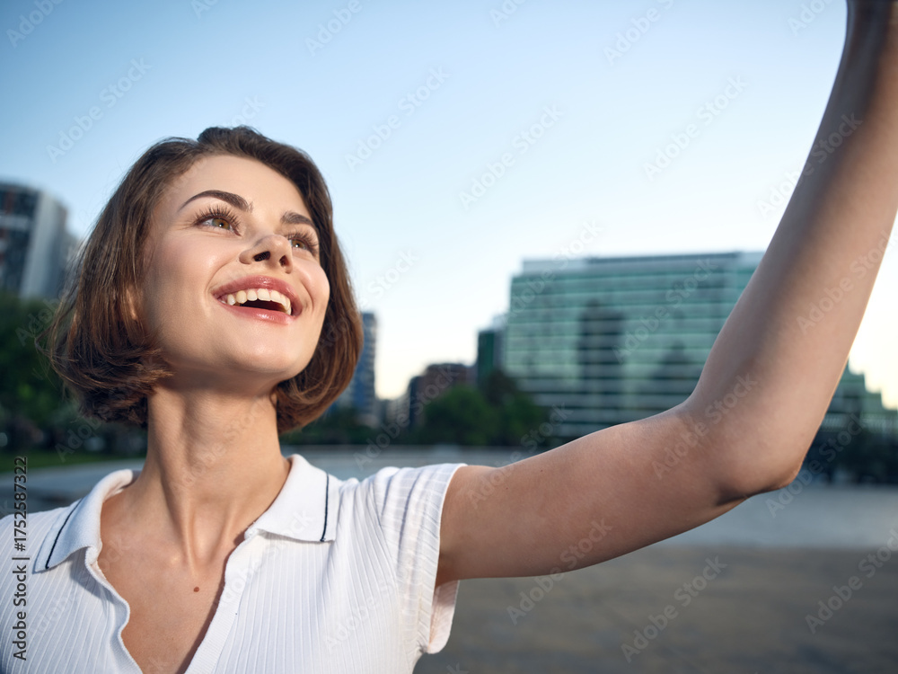 Fototapeta premium A cheerful young woman takes a selfie in a sunny urban park beside a river, smiling brightly as modern buildings rise in the background, conveying optimism, confidence, and a casual, upbeat lifestyle