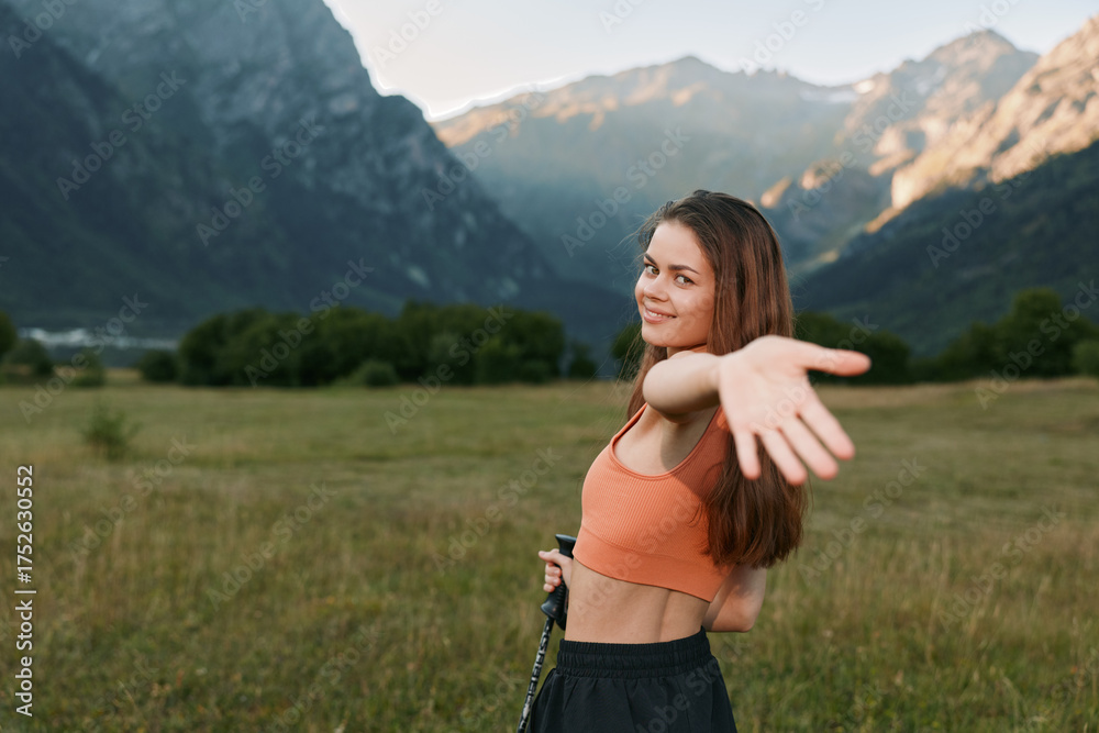 Obraz premium A cheerful young woman stands in a scenic valley, wearing a bright orange crop top. She smiles warmly and extends her arm toward the camera, creating a friendly, carefree outdoor mood.