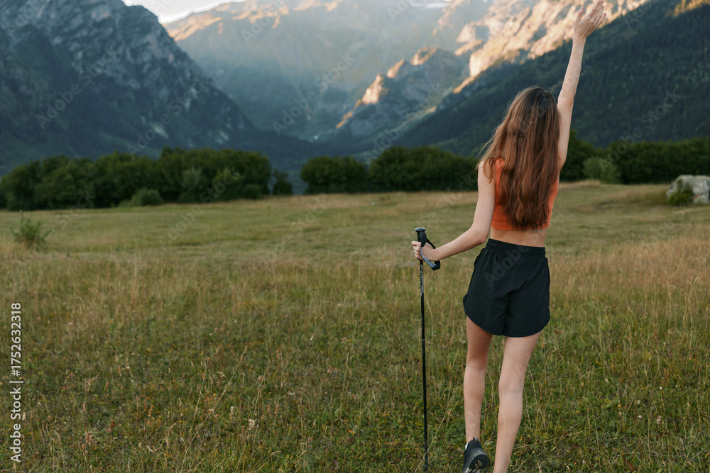 Fototapeta premium A young hiker stands in a grassy meadow with majestic mountains in the distance. She raises a hand in triumph, wearing a sleeveless top and shorts.