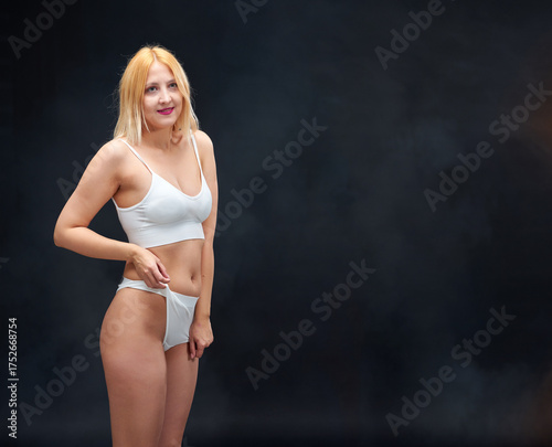 A young woman in white underwear is standing in a dark room. Her figure looks toned and athletic.