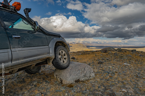 An off-road vehicle with mud tires is driving over a rock, with a view of the blue sky and mountains. A car tire is visible on the rock in the wild.