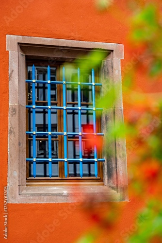 Farbenfrohes Fenster mit blauem Gitter an orangefarbener Fassade im Elsass – Detail historischer Architektur in Frankreich