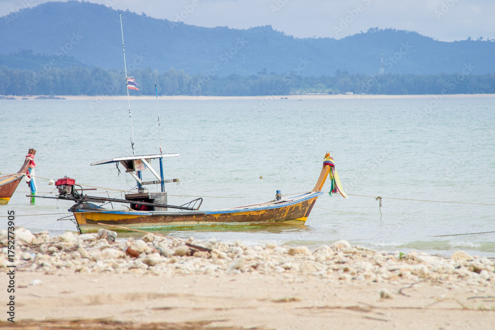 Naklejka premium Traditional Thai Fishing Boat Moored at a Rocky Tropical Shore. A colorful, classic longtail fishing boat anchored in the shallow water of a bay with rocky, sandy beach and distant forested mountains