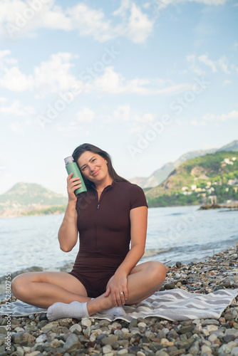 Healthy lifestyle concept. Woman sitting by the ocean, resting and hydrating after morning training.