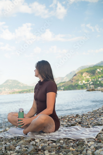 Morning workout by the sea. Fit woman relaxing on the beach and drinking water after exercise.