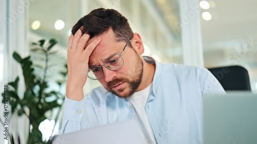 Worried businessman, dressed in a light blue shirt, processes upsetting news from a document in a bright office. Stressed specialist expresses frustration and disappointment with financial results.