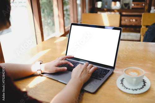 A person working on a laptop with a blank screen at a sunlit wooden table in a cafe, enjoying a cup of coffee. A concept for remote work or freelancing.