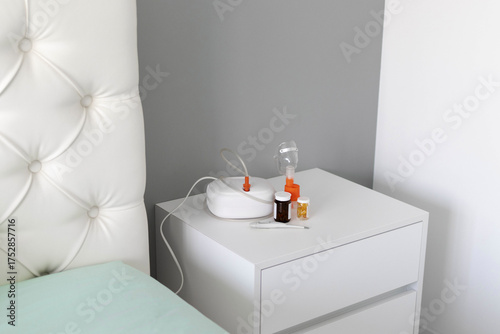 Close-up of a medical nebulizer with an inhalation mask, digital thermometer, and medicine bottles on the bedside table. Concept of respiratory therapy, healthcare at home, and asthma treatment.
