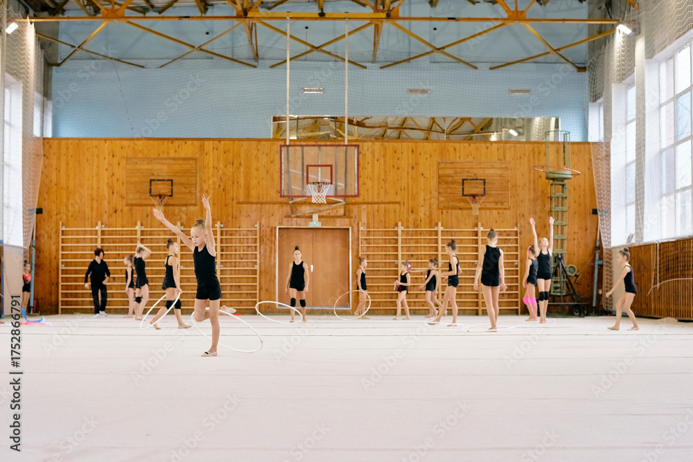Obraz premium Group of Caucasian girls practicing rhythmic gymnastics with hoops in sports hall, performing synchronized movements on gymnasium floor, some raising arms, others spinning hoops