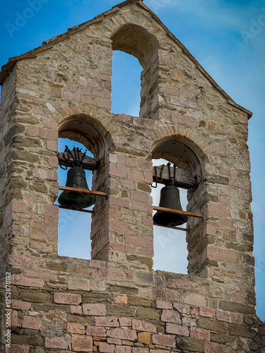 A close-up of a weathered, ancient stone bell tower featuring two dark bronze bells framed by rustic arches against a bright blue sky.