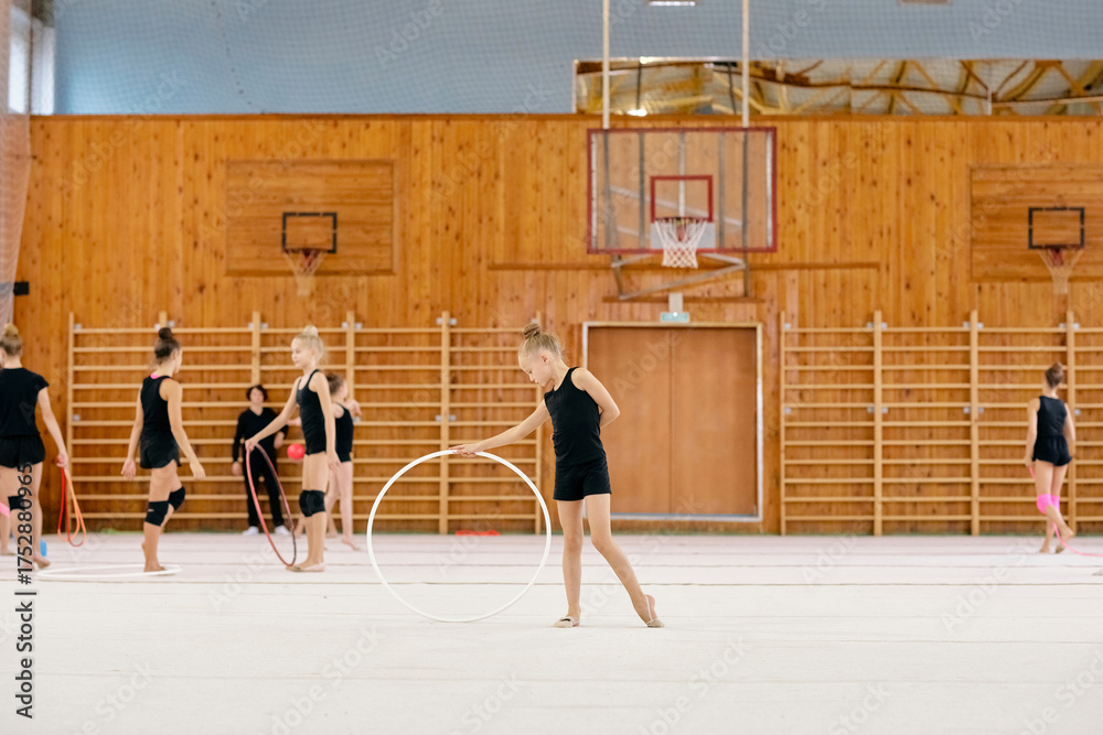 Obraz premium Caucasian girls practicing rhythmic gymnastics with hoops in gymnasium, young athletes performing exercises on mat, one girl standing in foreground holding hoop, others training behind
