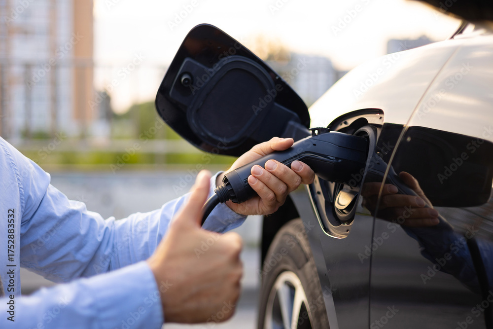 Fototapeta premium A person plugs in an electric vehicle charging cable, giving a thumbs up, symbolizing approval of the technology.