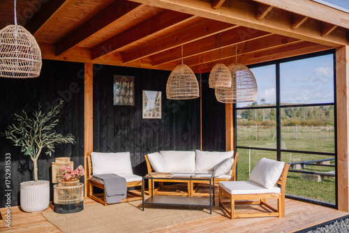 Stylish outdoor lounge with wooden furniture, rattan pendant lights, potted plants, and soft cushions set in a cozy covered patio with black wood walls.