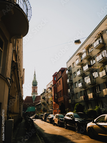 Parked cars lining narrow street in Krakow, Poland, with St. Florians Gate rising majestically in background, bathed in warm glow of late afternoon sun. Historic landmark. Cars parked along street