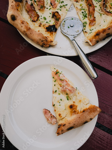 Slicing delicious pizza topped with crispy bacon and fresh chives on rustic wooden table, with tempting slice placed on white plate, ready for sharing. Slice of pizza with crispy bacon on white plate