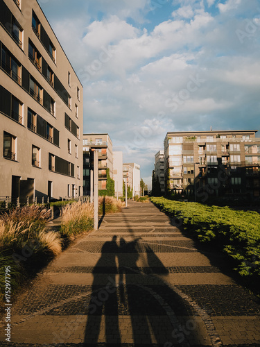 Long shadows of people walking on paved path in modern residential district with apartment buildings, bushes and scenical grasses under sky with clouds. Pedestrians casting shadows on urban walkway