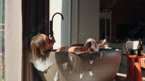 Smiling woman enjoys a relaxing bubble bath in a modern freestanding tub inside a rustic wooden cabin. The soft morning light creates a peaceful, intimate atmosphere. Concept of self-care, morning