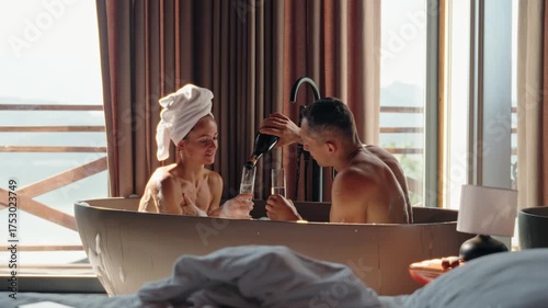 Romantic couple enjoying a hot bubble bath together while clinking champagne glasses in a rustic wooden cabin. Morning light pours in through the window, creating an intimate and serene atmosphere
