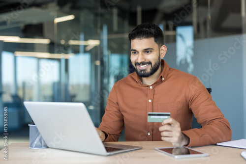 Smiling indian man sitting at a desk in a modern office, happily using a laptop and holding a credit card, engaging in convenient online shopping and e-commerce transactions
